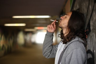 Auf dem Bild ist ein Jugendlicher zu sehen, der mit geschlossenen Augen, genüsslich einen Joint in einer Unterführung raucht. 