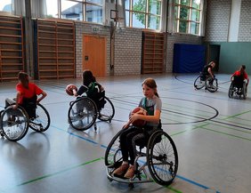 Auf dem Bild sind Kinder in Rollstühlen in einer Turnhalle zu sehen, die mit Basketbällen spielen.