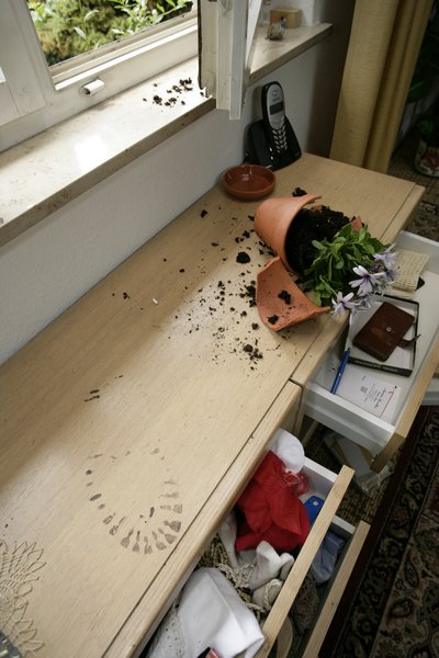 Auf dem Bild ist eine Kommode mit offenen, durchwühlten Schubladen zu sehen. Auf der Kommode liegt ein zerbrochener Blumentopf.