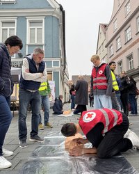 In der Fußgängerzone liegt eine Matte mit Übungspuppe auf dem Boden. Ein Schüler demonstriert Handgriffe an der Puppe, Passanten stehen drumherum und sind aufmerksam dabei.
